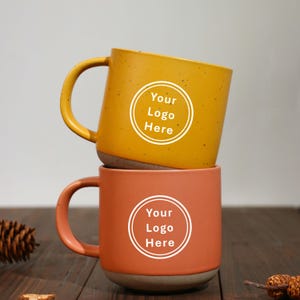 May include: Two ceramic mugs stacked on top of each other. The top mug is mustard yellow with a white circle that says "Your Logo Here". The bottom mug is orange with a white circle that says "Your Logo Here".