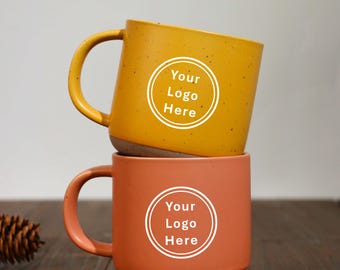 May include: Two ceramic mugs stacked on top of each other. The top mug is mustard yellow with a white circle that says "Your Logo Here". The bottom mug is orange with a white circle that says "Your Logo Here".