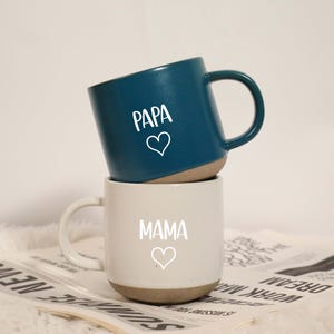 Può includere: Due tazze in ceramica impilate. La tazza superiore è verde acqua con la scritta "PAPA" e un cuore. La tazza inferiore è bianca con la scritta "MAMA" e un cuore. Entrambe hanno una base beige.