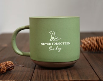 Puede incluir: Una taza de cerámica verde con base y asa marrón. La taza presenta un contorno blanco de un perro y el texto "NEVER FORGOTTEN Bailey". La taza está sobre una superficie de madera oscura.