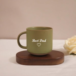 Könnte beinhalten: Eine grüne Keramiktasse mit den Worten "Best Dad" und einem Herzsymbol auf der Seite. Die Tasse steht auf einem kleinen, dunkelbraunen Holzuntersetzer. Neben der Tasse liegt eine cremefarbene Rose.