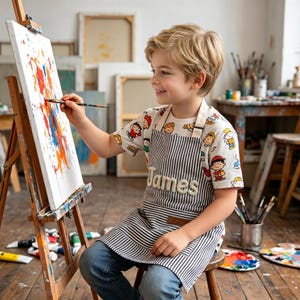 Puede incluir: Un joven artista con un delantal a rayas con el nombre "James" bordado y una camiseta de personajes de dibujos animados, pinta sobre un lienzo. El delantal es a rayas blancas y negras. El lienzo muestra salpicaduras de pintura coloridas.