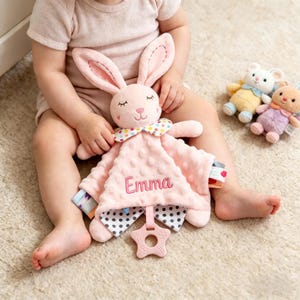 Puede incluir: Una manta de seguridad rosa para bebé con una cabeza de conejo y un mordedor en forma de estrella. La manta tiene bordado el nombre "Emma". También se ven dos pequeños peluches.