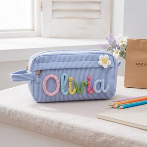 Puede incluir: Un estuche de pana azul claro con el nombre Olivia en letras trenzadas, coloridas y en relieve en la parte frontal. Una pequeña flor blanca de ganchillo con centro amarillo está fijada en la esquina superior derecha, con un asa lateral.