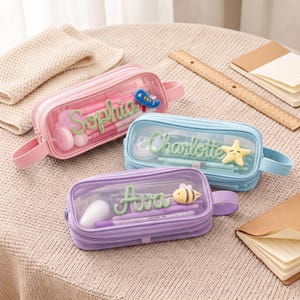 Puede incluir: Tres estuches rectangulares en rosa pastel, azul claro y lavanda, cada uno con un panel frontal transparente, un asa lateral y un nombre personalizado en letra cursiva verde con un pequeño icono de peluche de avión, estrella o abeja.