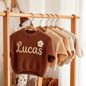 Puede incluir: Una fila de suéteres infantiles en varios colores neutros cuelgan de un estante de madera. El suéter delantero es de punto color óxido con el nombre "Lucas" bordado en crema y una aplicación de margarita.