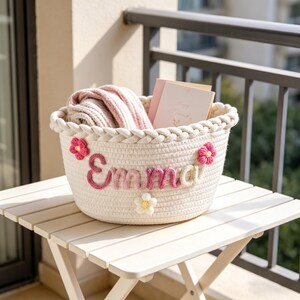 May include: A white woven basket with the name "Emma" in pink yarn letters, decorated with pink and white flower accents. The basket contains a folded pink blanket and a greeting card. It sits on a white folding table.