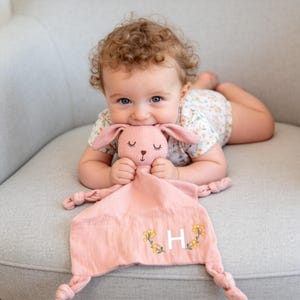 Puede incluir: Una manta de seguridad rosa con forma de conejito con bordado floral y la letra "H". La manta tiene una base anudada y la sostiene un bebé con un body floral. El conejito tiene los ojos cerrados y orejas largas.
