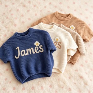 May include: Three short-sleeved knit sweaters in blue, cream, and tan. Each sweater features a ribbed crew neckline and a textured, embroidered daisy flower above the name James in a cursive, raised white or brown font on the front chest.