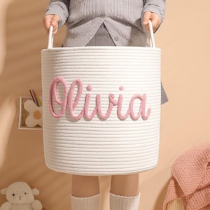 May include: A white rope basket with the name "Olivia" in pink script. The basket has two white handles and is held by a person. The basket is cylindrical and appears to be for storage.