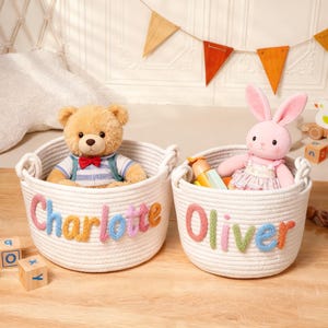 Puede incluir: Dos cestas de cuerda trenzada blanca con asas integradas. La cesta izquierda muestra el nombre Charlotte en letras de lana de colores y contiene un oso de peluche beige. La cesta derecha muestra el nombre Oliver en letras de lana de colores y contiene un conejo de peluche rosa.