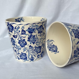 Puede incluir: Dos tazas de cerámica con base color crema y un estampado floral azul. Una taza está en posición vertical, la otra está de lado. El diseño floral azul cubre el exterior de las tazas, con un borde festoneado en la parte superior.