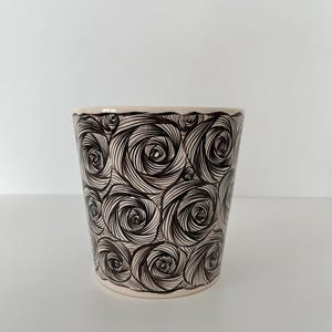 Puede incluir: Una maceta de cerámica con base color crema y un patrón repetido de diseños de rosas negras en espiral. La maceta tiene una forma ligeramente acampanada y un acabado liso y brillante. El diseño cubre todo el exterior de la maceta.