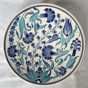 Peut inclure: Une assiette ronde en céramique avec un fond blanc et un motif floral bleu. Les fleurs sont dans des tons de bleu et de turquoise, avec des détails complexes. L'assiette a une bordure bleu foncé.