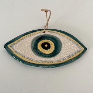 May include: Ceramic eye-shaped wall hanging with a textured off-white iris, surrounded by a teal and yellow border. The pupil is black, and the eye has a jute string for hanging. A decorative piece.