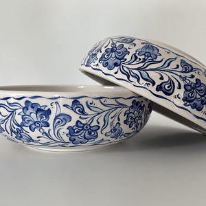 May include: Two ceramic bowls with a cream-colored base and a blue floral design. The bowls are decorated with intricate blue flowers and vines. One bowl is upright, and the other is tilted, revealing the interior design.