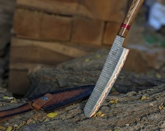 May include: A kitchen knife with a patterned Damascus steel blade featuring a wavy copper-coloured edge, a wooden handle with a red accent ring, and a matching dark brown leather sheath with a silver-coloured metal snap button closure.
