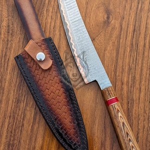 May include: A handcrafted knife with a wooden handle and a patterned blade, next to a leather sheath. The handle has a red accent. The sheath is brown with a basketweave design and a snap closure.