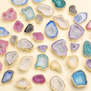Puede incluir: Una colección de colgantes de geoda coloridos con bordes dorados. Los colgantes presentan varios tonos de rosa, azul, morado y verde, con un interior cristalino. Cada colgante tiene un pequeño lazo para sujetar a una cadena.