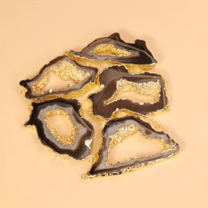 May include: A collection of five irregular agate slices featuring dark brown and grey banded patterns with central crystalline geode cavities. Each slice is framed by a metallic gold electroplated edge and includes small gold jump ring loops.