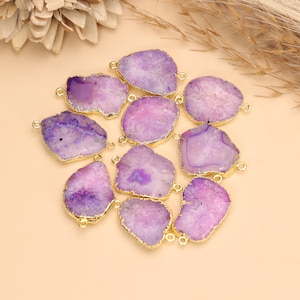 May include: A collection of ten irregular, purple agate stone slices, each featuring a gold-plated metallic edge and two small circular loops on opposite sides for connecting, arranged in a cluster on a light beige surface.