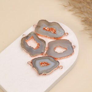 May include: Four irregular gray agate slices with rose gold electroplated edges. Each stone features a central geode cavity with sparkling crystals and two small metal loops attached to the sides for use as jewelry connectors or pendants.