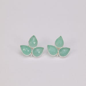 May include: A pair of silver-toned stud earrings, each featuring three light green, teardrop-shaped gemstones arranged in a leaf-like design. The gemstones have a faceted cut, and the earrings are set against a plain white background.