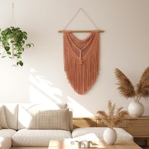 May include: A bright living room with a neutral colour scheme. A cream-coloured sectional sofa, a wooden coffee table, and a jute rug are in the centre. A macrame wall hanging and hanging plants add texture to the space.