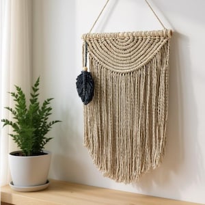 May include: A beige macrame wall hanging with a half-circle design and long fringe. A black feather with a wooden bead hangs from the side. The wall decor is suspended from a natural rope.