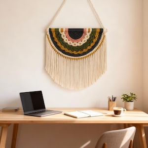 Könnte beinhalten: Ein Makramee-Wandbehang mit einem Halbkreis-Design in Schwarz-, Rosa-, Grün- und Goldtönen hängt über einem Holztisch. Auf dem Tisch befinden sich ein Laptop, ein Notizbuch, Bleistifte und eine Kaffeetasse. Eine kleine Pflanze steht daneben.