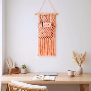 以下が含まれることがあります： 木製のダボとフリンジが付いた、桃色のマクラメの壁掛け。織り込まれたポケットには、ノートとペンが入っています。壁掛けは、ラップトップ、ノート、マグカップのある木製の机の上にあります。