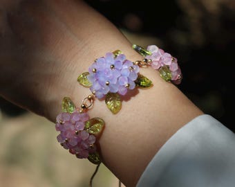Puede incluir: Una delicada pulsera con racimos de cuentas de flores lavanda y rosa con detalles dorados y hojas verdes. El diseño floral está conectado por una cadena dorada, creando un accesorio elegante.