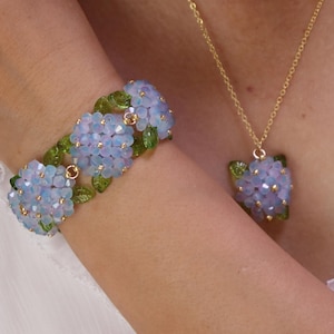 May include: A bracelet and a necklace set featuring light blue, faceted, flower-shaped beads arranged in clusters. Each cluster is accented with small, green, leaf-shaped beads and gold-toned metal connectors. The necklace hangs on a gold-toned chain.