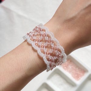 Könnte beinhalten: Ein breites Perlenarmband mit einem Gittermuster aus durchscheinenden, hellrosa facettierten Kristallen, eingefasst von gewellten Rändern aus kleinen, undurchsichtigen weißen Saatperlen, das ein zartes und komplexes gewebtes Design bildet.