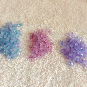 May include: Three piles of faceted beads in shades of blue, pink, and purple. The beads are scattered on a textured, off-white surface. The beads appear to be made of glass or a similar material, reflecting light.