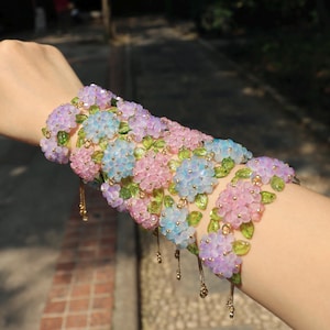 Könnte beinhalten: Ein Blumenarmband mit rosa, blauen und lila Perlenclustern, akzentuiert mit grünen, blattförmigen Perlen. Das Armband hat goldfarbene Kettenanhänger. Der Schmuck ist für das Handgelenk konzipiert.