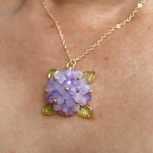 May include: A gold necklace with a pendant crafted to resemble a hydrangea flower. The pendant is made of lavender and purple beads, with green leaf accents. The necklace hangs from a fine gold chain.