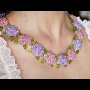 May include: A necklace with clusters of pink and lilac hydrangea flowers. The necklace has small green leaf accents and gold-coloured chain links. The floral design is set against a white background.
