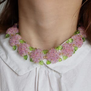 May include: A pink beaded necklace with flower-shaped clusters and green leaf details. The necklace is worn over a white collared shirt. The necklace is a delicate piece of jewellery.
