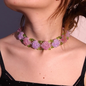 Puede incluir: Un collar choker floral con racimos de cuentas rosas y moradas, intercaladas con cuentas verdes en forma de hojas. El collar se lleva alrededor del cuello, mostrando su delicado diseño y colores vibrantes.