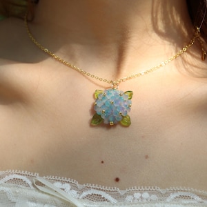 Puede incluir: Un delicado collar dorado con un colgante en forma de flor de hortensia. El colgante tiene cuentas azules claras y blancas con detalles de hojas verdes. El collar se lleva alrededor del cuello.