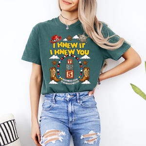 May include: A dark green short-sleeve t-shirt featuring a graphic print with the text I KNEW IT I KNEW YOU. The design includes a red cowboy hat, a sheriff star, two brown cowboy boots, and a central stack of three blocks labeled T, S, and 5.