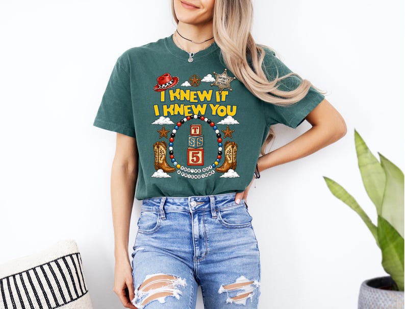 May include: A dark green short-sleeve t-shirt featuring a graphic print with the text I KNEW IT I KNEW YOU. The design includes a red cowboy hat, a sheriff star, two brown cowboy boots, and a central stack of three blocks labeled T, S, and 5.
