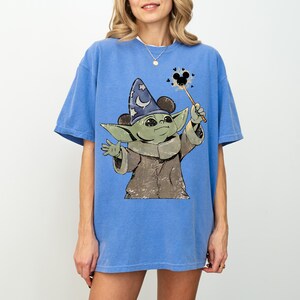 May include: A blue short-sleeve t-shirt featuring a graphic of a green creature wearing a blue wizard hat with stars and a crescent moon. The creature holds a wand with a black mouse-ear icon at the tip, surrounded by small black hearts.