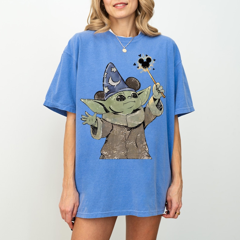 May include: A blue short-sleeve t-shirt featuring a graphic of a green creature wearing a blue wizard hat with stars and a crescent moon. The creature holds a wand with a black mouse-ear icon at the tip, surrounded by small black hearts.