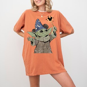 May include: An orange short-sleeve t-shirt featuring a graphic of a green creature wearing a blue wizard hat with white stars and moons. The creature holds a wand with a Mickey Mouse icon at the tip, surrounded by small black heart shapes.