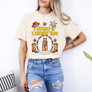 May include: A cream-colored t-shirt featuring a graphic print with the text I KNEW IT I KNEW YOU, a red cowboy hat, a sheriff star, cowboy boots, and a central stack of alphabet blocks labeled T, S, and 5, all surrounded by a colorful bead border.