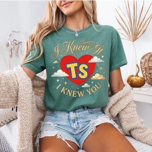 May include: A green short-sleeve t-shirt featuring a red heart graphic with the yellow letters TS in the centre. The text I KNEW IT and I KNEW YOU is printed in gold above and below the heart, surrounded by small gold stars and white clouds.