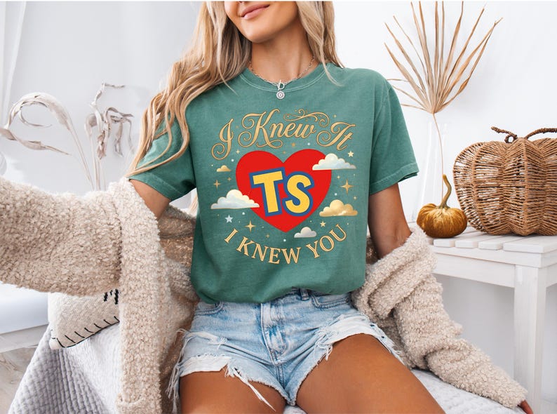 May include: A green short-sleeve t-shirt featuring a red heart graphic with the yellow letters TS in the centre. The text I KNEW IT and I KNEW YOU is printed in gold above and below the heart, surrounded by small gold stars and white clouds.