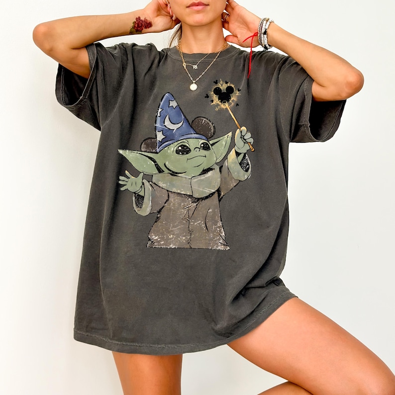 May include: A charcoal gray short-sleeve t-shirt featuring a graphic print of a green creature wearing a blue wizard hat with a crescent moon and stars. The creature holds a wand with a glowing Mickey Mouse icon at the tip, rendered in a distressed style.